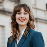 Lily Smogor poses in front of a white building background, smiling with red lipstick and wears a blue blazer and a bolo tie