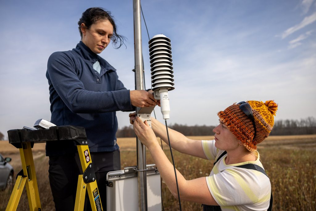 A temperature and humidity sensor is installed by two individuals in a rural area on a sunny day with a blue sky in the background.