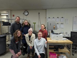 Six individuals pose together in a kitchen workspace, smiling.
