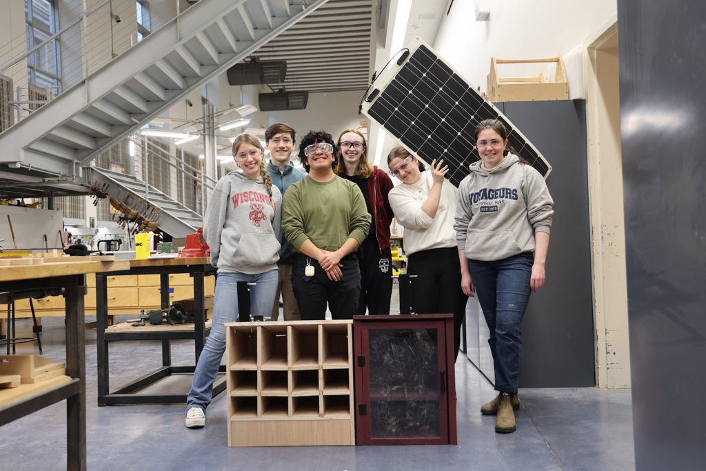 Students gather around a little free library, smiling and holding up a solar panel.