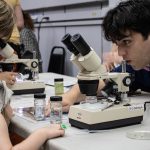 A CFL undergrad is leaning over a table helping a young person with a microscope during an Open House.