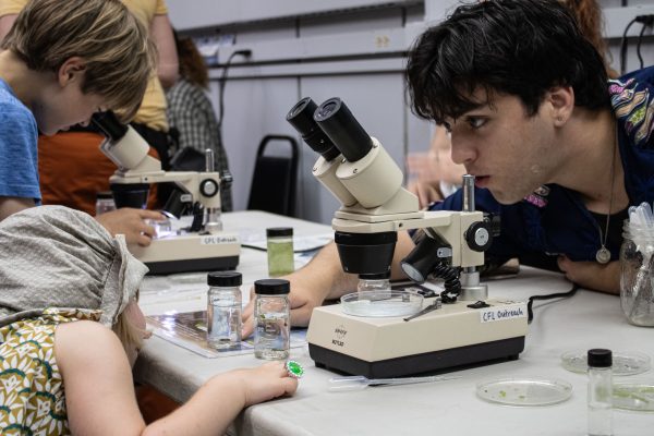 A CFL undergrad is leaning over a table helping a young person with a microscope during an Open House.