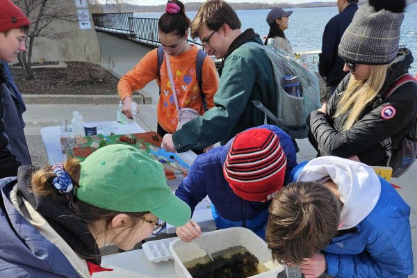 Group of people are standing outside of Hasler Lab with Lake Mendota in background. They are wearing colorful coats and hats as the weather is cool. They are peering at aquatic plant life in containers.