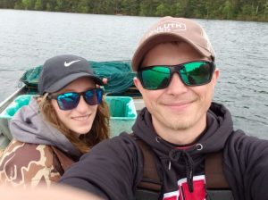 Joe is in the foreground, Amber is behind him and they are in a boat on a lake. They are smiling at the camera and wearing sunglasses.