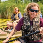 Carrie is in the foreground, wearing a camoflauge life jacket, holding a kayak padle, and smiling at the camera