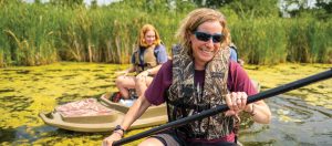Carrie is in the foreground, wearing a camoflauge life jacket, holding a kayak padle, and smiling at the camera