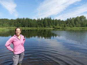 Katie has her hands on her hips and is standing beside of a calm lake with tree line in the background