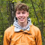 Steven is wearing an orange rain coat and is looking at the camera and smiling. He has thick curly brown hair and there are trees in the background.
