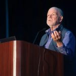 Steve Carpenter is standing at a podium, looking out at audience. He is holding his left hand up in gesture and is speaking to the audience.