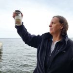 Emily Stanley is standing by the CFL dock, she's holding a jar of Lake Mendota water up and is looking up at it. Lake Mendota is in the background.