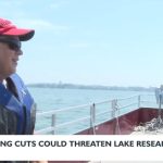 Emily Stanley is being interviewed on CFL pontoon boat. She is wearing a UW red ball cap, life jacket and sun glasses and looking to the side. The words "Federal Funding Cuts Could Threaten Lake Research" New 3 Now are part of the image.
