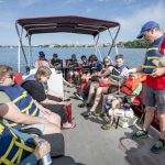 A group of people are sitting on CFL pontoon boat listening to Adam talk about one of the ways lakes are sampled.