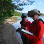 Two women wearing hats, sunglasses and red clothing are standing on shore of lake. One is holding a clipboard and a pencil recording information and both are looking down at the paper.