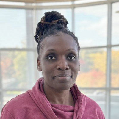 African American woman with her hair tied up in a bun. Smiling at camera, and wearing a pink sweatshirt. Picture taken in front of windows.