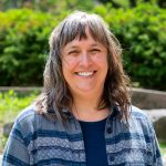 Picture of a white woman with wavy grey/brown hair and bangs. Smiling at the camera and standing outside.