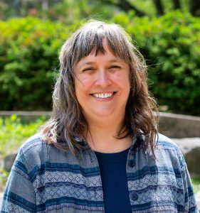 Picture of a white woman with wavy grey/brown hair and bangs. Smiling at the camera and standing outside.