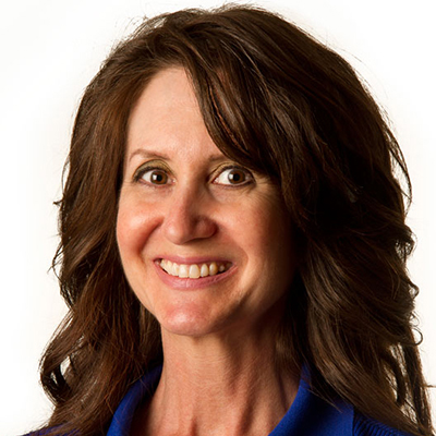 Woman with dark brown hair smiling at camera. In front of white background. Wearing a blue collared shirt.
