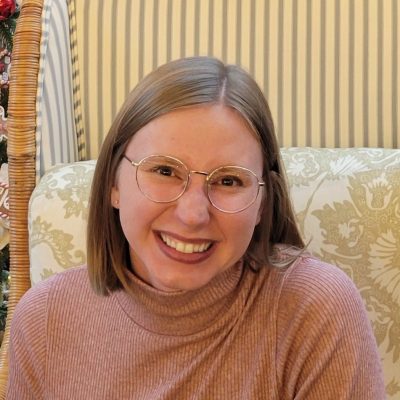 Young light brown-haired white woman smiling with metal glasses and short, light brown hair. She is wearing a pink top.