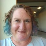 Middle-aged white woman with short brown hair. The ends of her hair are dyed blue and purple. She is wearing a white top.