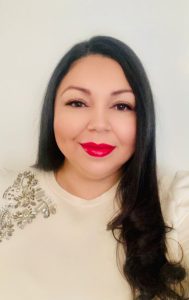 Photo of a hispanic woman with long dark hair. Wearing a cream top. Bright pink lipstick.