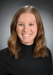 Photo of a young woman with shoulder-length brown hair. Wearing a black turtleneck.