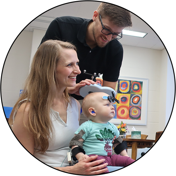 An infant participant seated in her mother’s lap. The infant has a 3-pointed head tracker on her forehead that allows the neuronavigation system to track her head. A smiling TMS operator holds a TMS coil above her head.