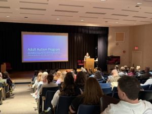 Madeline Barger presents the Adult Autism Program at the Day with the Experts: Autism. Madeline is at the podium in front of a power point presentation that reads Adult Autism Program. There is a full auditorium.