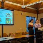 Monica and Jacqueline standing in front of a podium. Screen behind them with images and text in Spanish