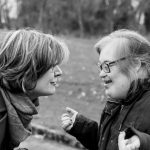 Woman with short hair smiling and looking at a young woman with Down syndrome, who is smiling and looking at the woman with short hair. Grass and trees in the background.